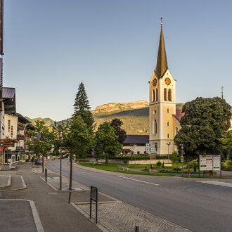 © Kleinwalsertal Tourismus | Steffen Berschin Eine ruhige Straße in einem charmanten Dorf mit einer hohen Kirche im Hintergrund. Umgeben von Bäumen und Bergen strahlt die Szene Ruhe aus. | © Kleinwalsertal Tourismus | Steffen Berschin