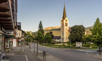 © Kleinwalsertal Tourismus | Steffen Berschin Eine ruhige Straße in einem charmanten Dorf mit einer hohen Kirche im Hintergrund. Umgeben von Bäumen und Bergen strahlt die Szene Ruhe aus. | © Kleinwalsertal Tourismus | Steffen Berschin