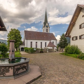 © Kleinwalsertal Tourismus | Steffen Berschin A picturesque village square with a fountain in the foreground and traditional houses. In the background, there is a church with a tall tower and a cloudy sky. | © Kleinwalsertal Tourismus | Steffen Berschin