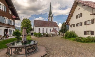 © Kleinwalsertal Tourismus | Steffen Berschin Ein malerischer Dorfplatz mit einem Brunnen im Vordergrund und traditionellen Häusern. Im Hintergrund ist eine Kirche mit einem hohen Turm und einem bewölkten Himmel zu sehen. | © Kleinwalsertal Tourismus | Steffen Berschin