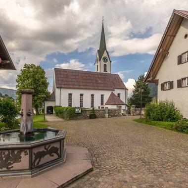 Ein malerischer Dorfplatz mit einem Brunnen im Vordergrund und traditionellen Häusern. Im Hintergrund ist eine Kirche mit einem hohen Turm und einem bewölkten Himmel zu sehen. | © Kleinwalsertal Tourismus | Steffen Berschin