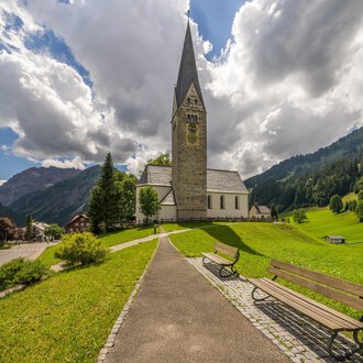 A picturesque church with a pointed tower stands in a green landscape. In the background, gentle hills and clouds in the sky can be seen. | © Kleinwalsertal Tourismus | Steffen Berschin