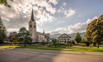A picturesque church with a tall tower stands next to a charming building. The sky is partly cloudy and the surroundings are green and inviting. | © Kleinwalsertal Tourismus eGen | Steffen Berschin