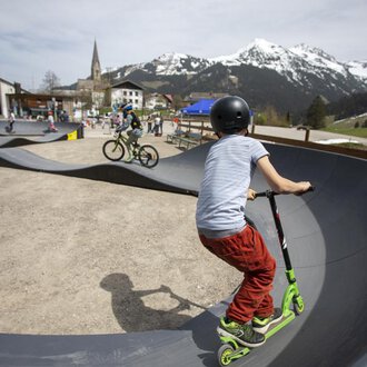 A skate park with children riding a scooter and a bike. In the background, mountains and a small town can be seen. | © Kleinwalsertal Tourismus | Frank Drechsel
