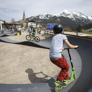 Ein Skatepark mit Kindern, die auf einem Roller und Fahrrad fahren. Im Hintergrund sind Berge und eine kleine Stadt zu sehen. | © Kleinwalsertal Tourismus | Frank Drechsel