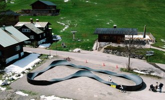 A modern, curved toboggan run in a picturesque mountain landscape. In the background, rustic wooden houses and green meadows can be seen. | © Christian Posch | Klaus Zopf