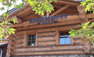 © Kleinwalsertal Tourismus | Veronika Senn Ein rustikales Holzgebäude mit einem Schild, das "Mühlenkammer" zeigt. Umgeben von grünen Blättern und einem klaren Himmel. | © Kleinwalsertal Tourismus | Veronika Senn