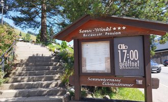 A wooden sign of the restaurant "Sonu-Winkl" displays the opening hours. In the background, a staircase and trees can be seen. | © Kleinwalsertal Tourismus | N. Lughammer