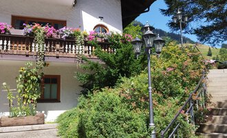 A beautiful house with blooming balconies and green patios. The staircase leads to a well-kept garden outdoors. | © Kleinwalsertal Tourismus | N. Lughammer