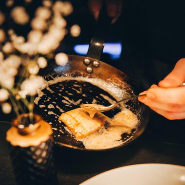A hand serves a golden-brown dish from a pan. In the background, there is a small flower vase.