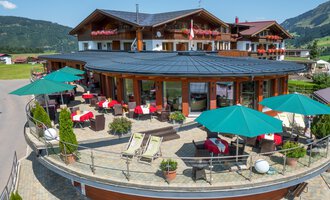 Ein modernes Hotel Birkenhöhe mit Terrasse, Sonnenschirmen und gemütlichen Sitzgelegenheiten. Im Hintergrund sind grüne Hügel und bergige Landschaften sichtbar.