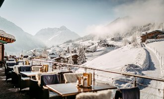 Eine gemütliche Sonnenterrasse im Hotel Birkenhöhe mit Tischen und Stühlen, umgeben von schneebedeckten Bergen. Der Himmel ist klar und die Landschaft ist malerisch.