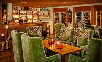 Eine gemütliche Lobby und Bar mit grünen Sesseln und einem Holztisch. Im Hintergrund sind Regale mit Spirituosen wie Rum, Whiskeyflaschen.