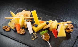 A cheese platter with various types of cheese, nuts, and fresh fruits. There is also a spoon with a fruity jam on a black plate. | © Hotel Alte Krone