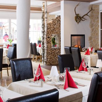 An elegant restaurant with modern tables and chairs. The tablecloths are cream-colored and the napkins are in red, while a fireplace in the background creates a cozy atmosphere. | © Aparthotel Kleinwalsertal Haller GmbH & Co KG