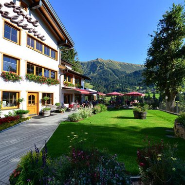 Ein schön gestaltetes Haus mit Balkonen und blühenden Blumen. Im Hintergrund sind grüne Berge und ein klarer Himmel zu sehen. | © Hotel Bellevue | I. Malouvier