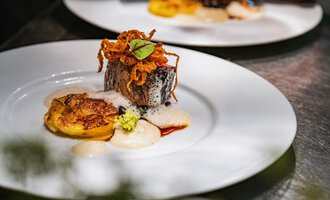 Ein kunstvoll angerichteter Teller mit gebratenem Fisch, knusprigen Zwiebeln und einer Beilage aus gebratenen Bananen. Dazu eine cremige Sauce und frische Kräuter. | © Verwöhn- & Wellnesshotel Walserhof| Frank Simon