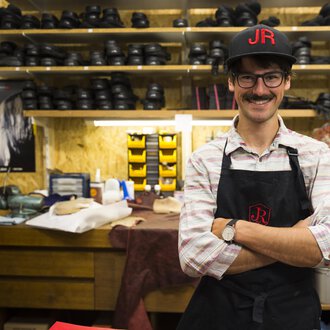 A cobbler stands in his workshop with a broad smile. In the background, many shoes and tools are visible. | © Kleinwalsertal Tourismus | Oliver Farys