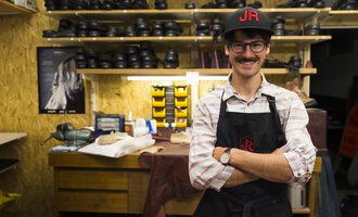 A cobbler stands in his workshop with a broad smile. In the background, many shoes and tools are visible. | © Kleinwalsertal Tourismus | Oliver Farys