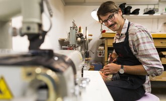A shoemaker in his workshop, working on a pair of shoes. Tools and machines are visible on the table. | © Kleinwalsertal Tourismus | Oliver Farys