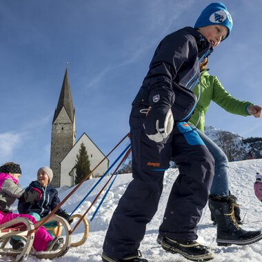Children are sledding in the snow while adults in warm clothes accompany them. In the background, there is a church and the blue sky is visible. | © Kleinwalsertal Tourismus | Dominik Berchtold