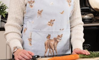 Eine Person trägt eine schicke Schürze mit Tiermotiven und steht in einer Küche. Vor ihr liegen frisches Gemüse auf einem Schneidebrett.