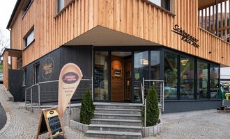 © Schönegger Käse-Alm Ein modernes Restaurant mit Holzfassade und großen Fenstern. Der Eingangsbereich ist einladend gestaltet, mit einem Schild für das Restaurant. | © Schönegger Käse-Alm