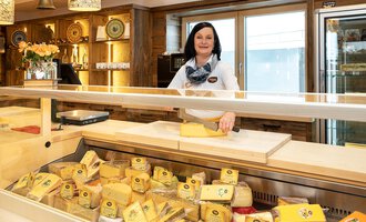 © Schönegger Käse-Alm Eine Käseverkäuferin steht hinter einer Theke voller Käsesorten. Der Laden hat ein gemütliches Ambiente mit Holzregalen und natürlichen Lichtquellen. | © Schönegger Käse-Alm