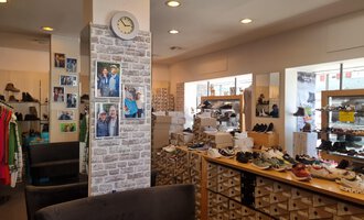 © Schuhhaus Schuster | Marietta Diehl Ein gemütliches Schuhgeschäft mit verschiedenen Schuhen und Bekleidung. An der Wand hängen Fotos, und eine Uhr ist sichtbar. | © Schuhhaus Schuster | Marietta Diehl