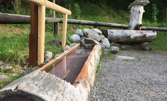 A rustic wooden fountain made from tree trunks in a natural setting. In the background, more wooden elements and vegetation can be seen. | © Kleinwalsertal Tourismus