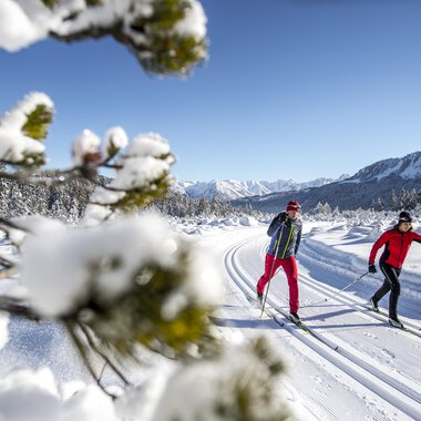 Two people are skiing on a snow-covered track. In the background, there are snow-capped mountains and a clear blue sky. | © Kleinwalsertal Tourismus | Dominik Berchtold