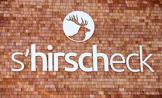 Ein Logo mit dem Namen "s'hirschcheck" ist auf einer Holzoberfläche angebracht. Das Design zeigt ein stilisiertes Geweih in einem runden Ausschnitt. | © Kleinwalsertal Tourismus | Frank Drechsel