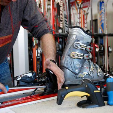 Ein Mann repariert Skiausrüstung und montiert einen Skischuh. Im Hintergrund stehen zahlreiche Ski.