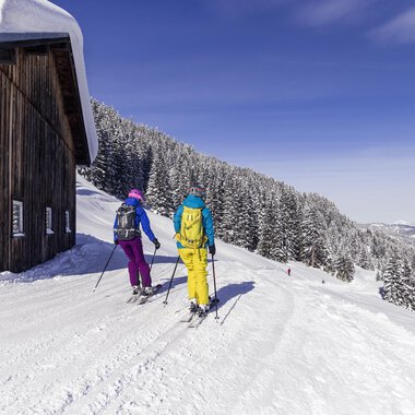 Zwei Skifahrer gehen einen schneebedeckten Weg entlang. Im Hintergrund sind Bäume und eine klare, blaue Himmel sichtbar. | © Kleinwalsertal Tourismus | Oliver Farys