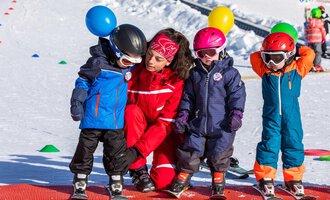 A ski instructor stands between four children in colorful winter clothing on a practice slope. The children are wearing helmets and are holding balloons in their hands. | © Skischule Bödmen-Baad GmbH