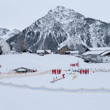 Eine verschneite Landschaft mit einem Dorf und einer Kirche im Vordergrund. Im Hintergrund ragen steile Berge empor, während eine Gruppe von Menschen in roten Jacken aktiv ist. | © Skischule Mittelberg | Karl Schuster