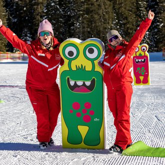 Two cheerful people in red suits are standing next to a colorful monster sign in the snow. In the background, trees and a snowy landscape can be seen.