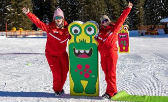 Two cheerful people in red suits are standing next to a colorful monster sign in the snow. In the background, trees and a snowy landscape can be seen.