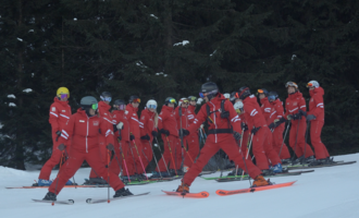 A group of skiers in red suits stands in a snowy ski area. The trees in the background create a winter atmosphere.