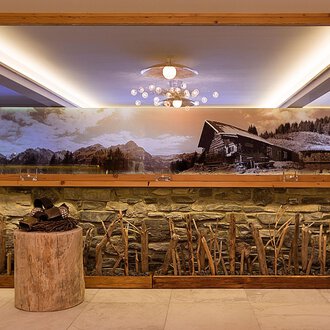 © Aparthotel Kleinwalsertal Haller GmbH & Co KG Ein elegantes Interieur mit einer Steinwand und einem großen Spiegel. Im Hintergrund ist ein großflächiges Landschaftsbild mit einer Hütte und Bergen zu sehen. | © Aparthotel Kleinwalsertal Haller GmbH & Co KG