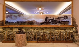 © Aparthotel Kleinwalsertal Haller GmbH & Co KG Ein elegantes Interieur mit einer Steinwand und einem großen Spiegel. Im Hintergrund ist ein großflächiges Landschaftsbild mit einer Hütte und Bergen zu sehen. | © Aparthotel Kleinwalsertal Haller GmbH & Co KG