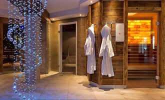 © Aparthotel Kleinwalsertal Haller GmbH & Co KG Ein modernes Wellness-Interior mit sanfter Beleuchtung. Links hängen weiße Bademäntel, und eine beeindruckende Wasserinstallation verleiht dem Raum ein entspannendes Ambiente. | © Aparthotel Kleinwalsertal Haller GmbH & Co KG
