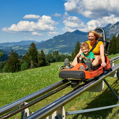 Ein glückliches Kind und eine Frau fahren auf einer Sommerrodelbahn. Im Hintergrund sind grüne Berge und ein blauer Himmel zu sehen. | © OBERSTDORF · KLEINWALSERTAL BERGBAHNEN