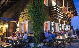A cozy wooden house with colorful windows and plants. Several people are sitting at long tables on it, enjoying the evening atmosphere. | © Sonna Alp | Wolfram Riezler