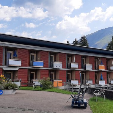 A modern building with multiple floors and balconies. The surroundings are green with a view of the mountains. | © Kleinwalsertal Tourismus | Veronika Senn