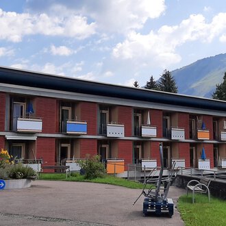 © Kleinwalsertal Tourismus | Veronika Senn A modern building with multiple floors and balconies. The surroundings are green with a view of the mountains. | © Kleinwalsertal Tourismus | Veronika Senn