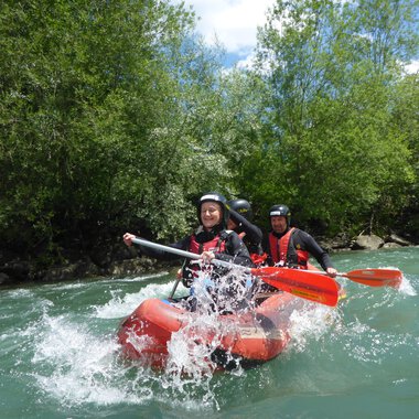 A group of people paddles on a red inflatable boat through fast currents. In the background, green trees and a blue sky can be seen. | © Spirits of Nature | Bernd Goller