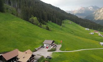 A tranquil landscape with green meadows and mountains in the background. In the foreground, several traditional huts and a pathway can be seen.