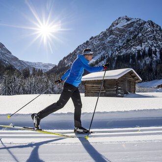 Ein Langläufer in blauer Kleidung fährt durch eine schneebedeckte Landschaft. Im Hintergrund sind Berge und eine kleine Hütte zu sehen, unter einem klaren blauen Himmel. | © Kleinwalsertal Tourismus | Dominik Berchtold