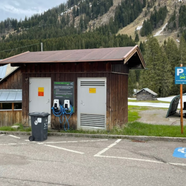 Eine kleine Holzgebäude mit Ladestationen für Elektroautos steht auf einem Parkplatz im Freien. Im Hintergrund sind grüne Wiesen und bewaldete Hügel zu sehen. | © Kleinwalsertal Tourismus